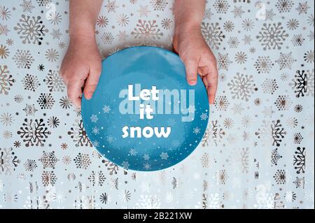 deux mains tenant une plaque bleue vide avec le décor des flocons de neige et l'inscription la laissent neiger sur un fond de papier d'emballage avec des flocons de neige brillants Banque D'Images