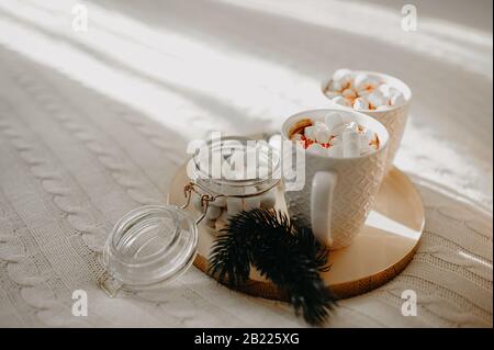 deux tasses blanches avec une boisson chaude et des guimauves blanches, sur un plateau doré avec un pot en verre de guimauves blanches sur une couverture tricotée blanche au soleil Banque D'Images