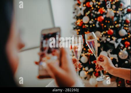 Fille fait une photo sur un téléphone blanc, deux verres de cheers avec champagne sur le fond de l'arbre de Noël dans les mains des femmes Banque D'Images