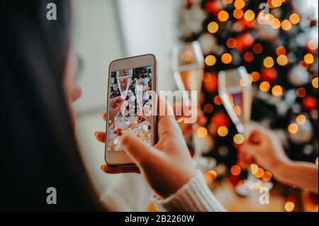 Fille fait une photo sur un téléphone blanc, deux verres de cognement avec du champagne sur le fond de l'arbre de Noël dans les mains des femmes Banque D'Images