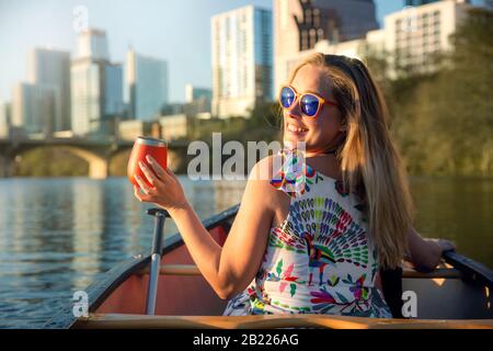 Jeune femme amusante avec des lunettes de soleil souriant, profitant de la vie et s'amuser en bateau sur une rivière à côté d'un horizon urbain Banque D'Images