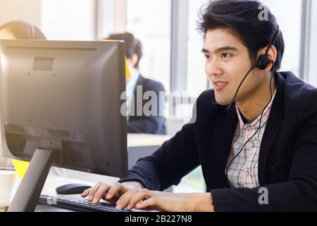 Un opérateur de centre d'appels asiatique souriant utilise un ordinateur pour communiquer avec le client ou effectuer une autre affectation. Banque D'Images
