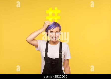 Hashtag, blogging. Portrait d'une jeune femme joyeuse avec des cheveux courts violets dans des salopettes en denim tenant un grand signe de hachage jaune sur sa tête et souriant à CAM Banque D'Images