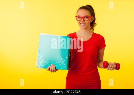 jolie jeune femme caucasienne heureuse dans un t-shirt rouge sur fond jaune vif geste puissance de la connaissance hodling un haltère et un grand dossier Banque D'Images