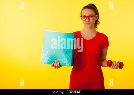 jolie jeune femme caucasienne heureuse dans un t-shirt rouge sur fond jaune vif geste puissance de la connaissance hodling un haltère et un grand dossier Banque D'Images