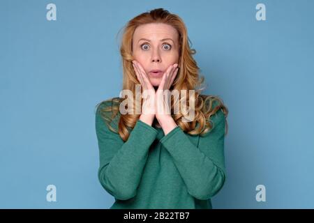 Choqué femme caucasienne mûre avec de longs cheveux debout sur fond bleu. La mère est dans la panique becaue que son enfant a fait une erreur. Studio tourné Banque D'Images