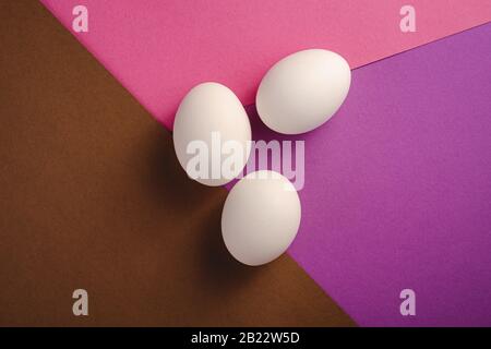 Trois oeufs blancs sur les couleurs pourpre, rose et brun fond simple minimal, vue de dessus, joyeuse journée de Pâques Banque D'Images
