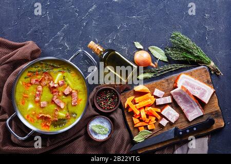 Soupe de pois fendus avec bacon, jambon de porc, carotte, pomme de terre au thym servie sur un pot avec feuille de laurier, sur fond de béton avec des ingrédients sur un boa de coupe Banque D'Images