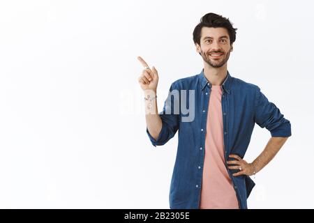 Un jeune homme gai plein de confiance avec un t-shirt rose, une chemise, tenez la main à la taille, pointant vers le coin supérieur gauche et souriant, suggérez à vos amis de se rendre à la fête Banque D'Images