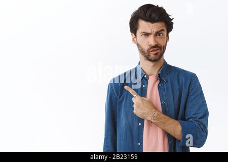 Sceptique, incertain et suspect, homme de 25 ans avec barbe, frognant smirk incertain, pointant vers la gauche et en affamant l'appareil photo douteux, ne peut pas décider Banque D'Images