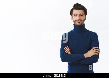 Entrepreneur masculin confiant et plein de confiance en pull bleu à col haut, bras croisés sur la poitrine, smirk assy et auto-assuré, affirmé debout. Beau Banque D'Images