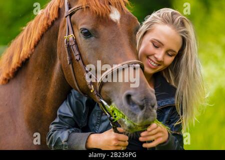 Belle femme souriante nourrissant son cheval arabe avec de l'herbe dans le champ Banque D'Images