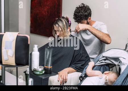 Maître coiffeur coupe les cheveux de femme blonde. Bébé dans le bébé alors que la mère a la coupe de cheveux dans le salon. Banque D'Images