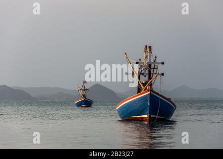 Deux petits bateaux de pêcheurs thaïlandais dans les couleurs nationales thaïlandaises ancrage rouge, blanc et bleu dans la mer avec quelques montagnes dans le fond lointain dans Banque D'Images