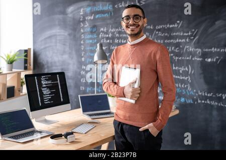 Jeune programmeur gai avec gadget mobile debout par table avec des ordinateurs portables et ordinateur avec tableau noir en arrière-plan Banque D'Images