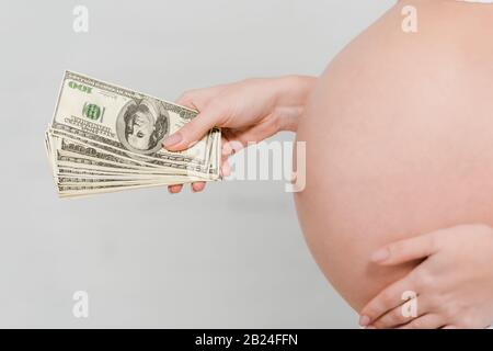 Vue rognée de la femme enceinte qui détient des billets en dollars sur fond gris, concept de maternité de mères porteuses Banque D'Images