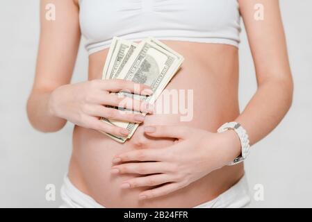 Vue rognée de la femme enceinte qui détient des billets en dollars près du ventre sur fond gris, concept de maternité de mères porteuses Banque D'Images