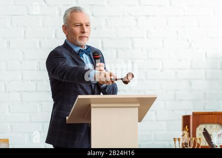 beau encanteur dans le costume tenant le microphone et pointant avec le gavel pendant la vente aux enchères Banque D'Images