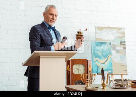 beau encanteur en costume pointant avec la main à la mouture de café et tenant le microphone pendant la vente aux enchères Banque D'Images