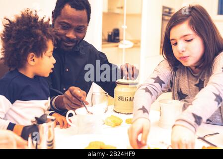 le papa noir à la table avec des enfants de race mixte ont le petit déjeuner, le papa aide l'enfant en versant du sucre dans sa tasse de lait, l'enfant sert le herse Banque D'Images