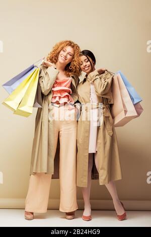 joyeuses filles multiculturelles en trench manteaux tenant des sacs de shopping sur beige Banque D'Images