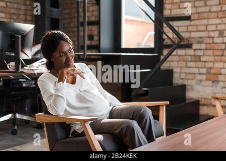 femme d'affaires afro-américaine souriante et enceinte assise dans un fauteuil et regardant loin Banque D'Images