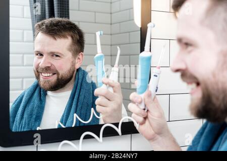 Un homme positif tient une brosse à dents rechargeable à la main et un irrigateur électrique et oral, regarde dans le miroir et sourit. Banque D'Images