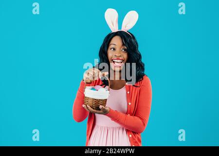 Portrait d'une mère excitée avec panier d'œufs de Pâques et oreilles de lapin regardant à côté, isolé sur fond bleu Banque D'Images