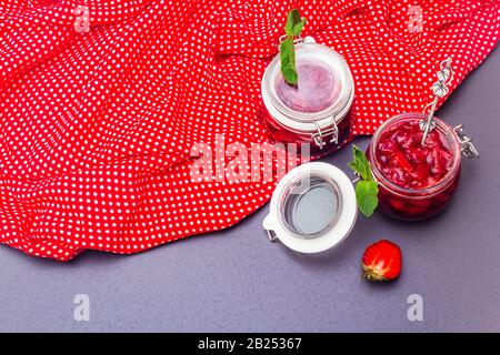 Confiture de fraises dans un pot en verre. Concept de préservation maison. Avec fraises fraîches et menthe, cuillère argentée sur fond de pierre noire, polka points clo Banque D'Images