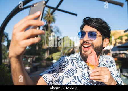 Portrait de jeune homme profitant du temps ensoleillé, prendre un selfie avec le téléphone et manger une glace à l'extérieur. Banque D'Images