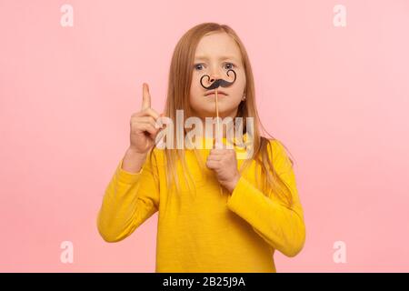 Bossy petite fille de gingembre avec des moustaches de papier drôle gestuant plus fin, donnant des conseils, prétendant être un homme intelligent, s'amuser avec mascarade accesso Banque D'Images