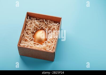 Oeuf en or dans une boîte en bois sur fond bleu. Le concept d'exclusivité et de superprix. Composition minimaliste. Vue de dessus, isolation Banque D'Images