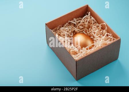 Oeuf en or dans une boîte en bois sur fond bleu. Le concept d'exclusivité et de superprix. Composition minimaliste. Vue de dessus, isolation Banque D'Images