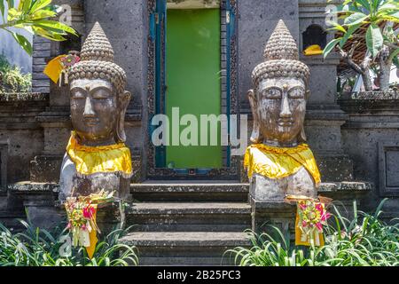 Statues en pierre de deux têtes de Bouddha décorées dans un tissu 'prada' doré près d'une maison balinaise traditionnelle à Desa Munggu, Bali, Indonésie. Banque D'Images