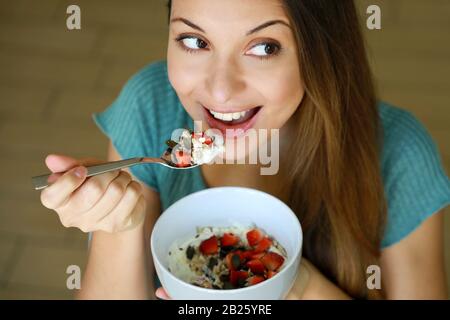 Vue rapprochée depuis le dessus de la belle jeune femme mangeant des yaourts skyr avec des fraises, des muesli de céréales et des graines à la maison, en regardant le côté, se concentrer sur Banque D'Images