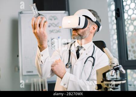 Homme mûr scientifique dans un laboratoire moderne lumineux, utilisant des lunettes de réalité virtuelle pour la recherche, en regardant la fiole en verre avec une nouvelle substance ou drogue. Vr Banque D'Images