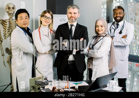 Groupe mixte de jeunes médecins, de travailleurs de laboratoire, de scientifiques, avec un homme de maturité masculin superviseur, travaillant dans le laboratoire moderne, souriant et tenant les bras croisés Banque D'Images