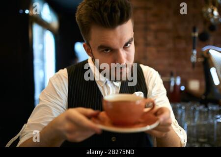 Tasse de barista sérieuse et élégante avec cappuccino à la lavande, demandant de goûter à la boisson .gros plan photo, tasse floue. Banque D'Images