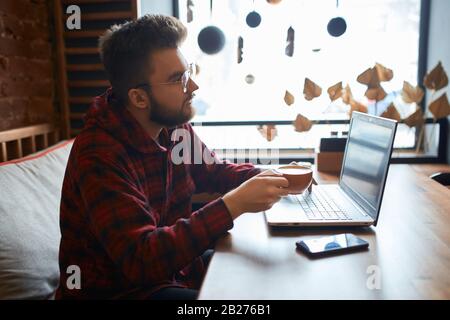 jeune homme dans des vêtements élégants et décontractés avec chat vidéo avec des amis, conversation en ligne, profiter d'un délicieux café à l'intérieur. vue latérale rapprochée ph Banque D'Images