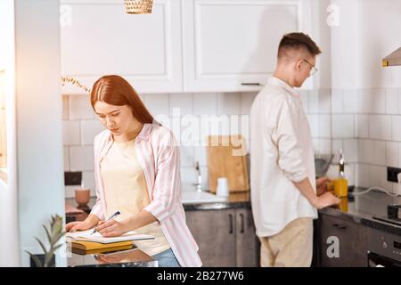 la jeune fille s'est concentrée sur la planification de sa journée, jeune homme dans une chemise blanche et un pantalon debout à côté de la cuisinière, tenant une tasse de thé. vie quotidienne Banque D'Images