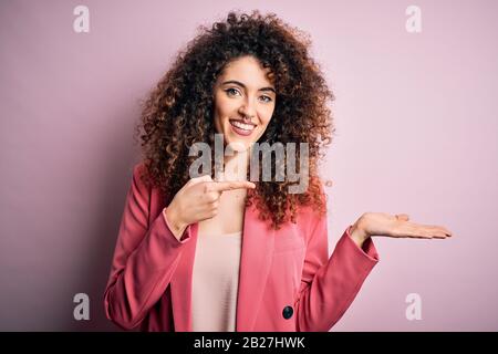Jeune femme d'affaires avec ses cheveux bouclés et piercing portant une élégante veste éblée et souriante à l'appareil photo tout en présentant avec la main et le poin Banque D'Images