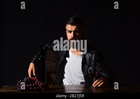 Degustator à visage confus est assis par une bouteille de vin et des raisins sombres. L'homme avec barbe tient un verre de vin sur fond brun foncé. Le sommelier a un goût de boisson chère. Concept de dégustation et de dégustation Banque D'Images