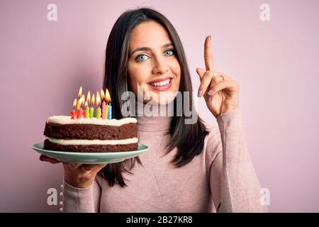 Jeune femme aux yeux bleus tenant un gâteau d'anniversaire avec des bougies sur fond rose surpris par une idée ou une question pointant doigt avec le visage heureux, Banque D'Images