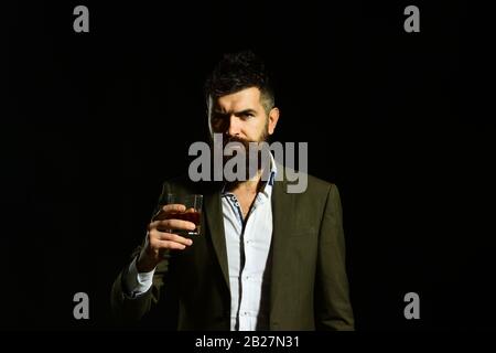 Homme au visage sérieux en costume tenant un verre d'alcool. Connaisseur avec boissons intelligentes cognac ou brandy. Concept de boisson et d'alcool. Homme d'affaires avec barbe et boissons sur fond noir Banque D'Images