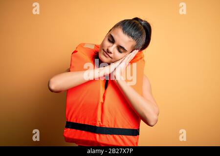 Jeune femme brunette portant un gilet de sauvetage orange sûr sur fond jaune dormant fatigué rêvant et se posant avec les mains ensemble tout en frottant Banque D'Images