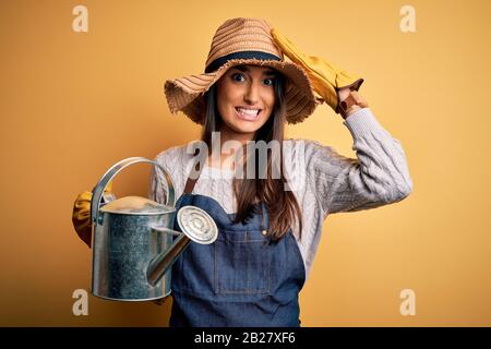 Une jeune femme agriculteur brunette portant un tablier et un chapeau tenant l'arrosage peut stressé avec la main sur la tête, choquée avec la honte et le visage surprise, ang Banque D'Images