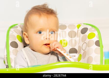 Très petit garçon avec un look bien pensé et une cuillère en plastique pour bébé dans la bouche s'asseoir dans la chaise haute Banque D'Images