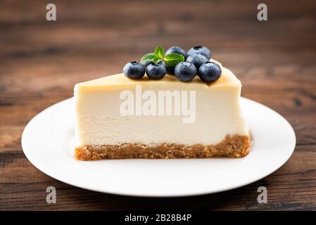Cheesecake Slice De New York Avec Des Bleuets Et De La Feuille De Menthe Sur La Plaque Blanche Fond De Table En Bois. Tranche Savoureuse De Gâteau Crémeux Banque D'Images