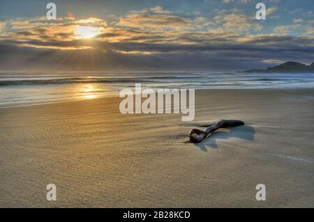 Orange Sun situé derrière les nuages qui encadrent l'horizon de l'océan Pacifique au large de la côte de l'Oregon, éclairant le sable de la plage et un seul morceau de dérivtwoo Banque D'Images