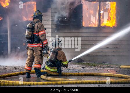 Deux pompiers en costumes de protection complets et réservoirs d'oxygène travaillant avec un long tuyau d'eau jaune cracher l'eau sur la maison brûlante fumage englouti dans le feu A Banque D'Images